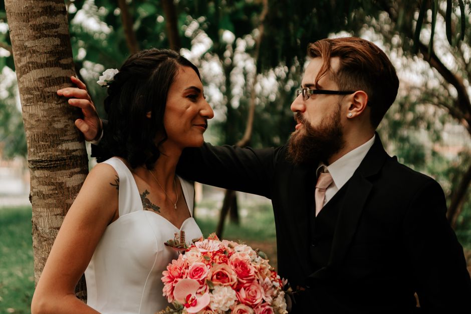 A bride and groom portrait outdoors in a park setting, with the bride holding a lush bouquet of pink roses, peonies, and greenery, featuring vibrant pink and soft peach hues. The bride is wearing a wh