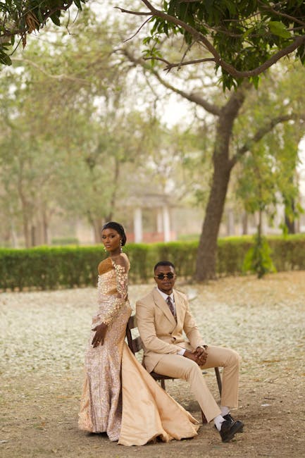 A woman in an elegant, off-the-shoulder lace wedding gown with intricate floral patterns and a full, flowing skirt stands beside a seated man in a beige suit, white shirt, and dark sunglasses. The sce