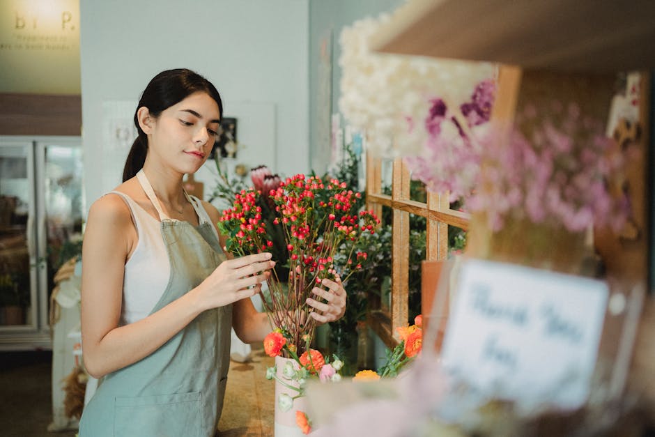 A young female florist with dark hair tied back, wearing a light-colored apron over a sleeveless top, arranging a vibrant bouquet of red and pink small, round flowers with green stems. She is standing
