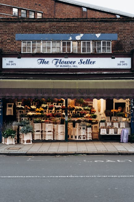 The exterior of The Flower Seller of Muswell Hill, a flower shop with a blue sign displaying contact information, sits beneath a weathered blue awning. The shop showcases a vibrant floral display, fea