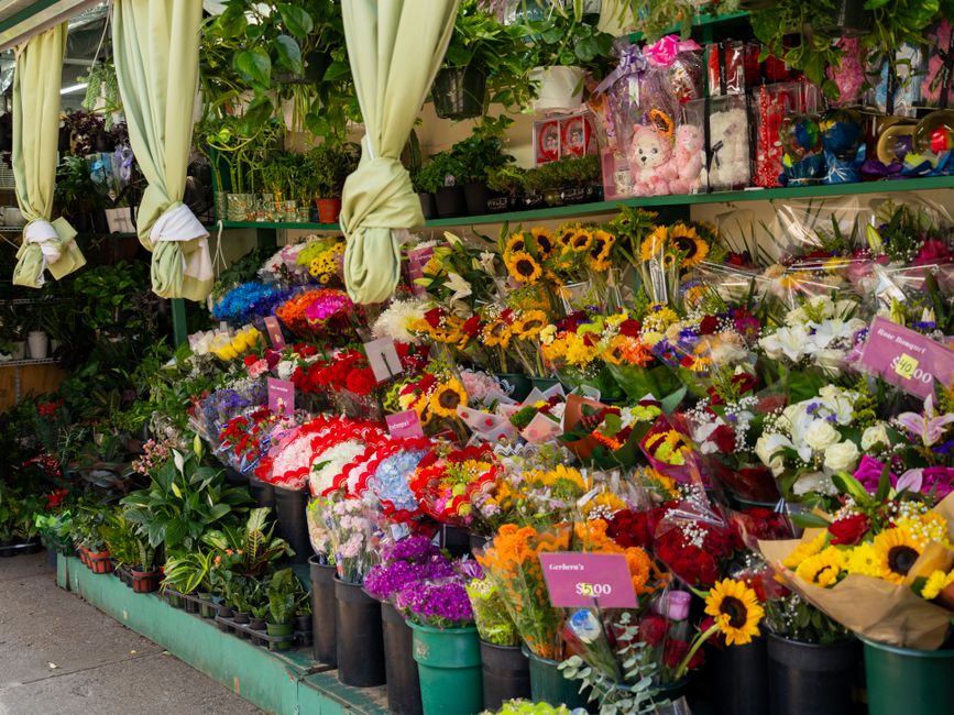 A vibrant outdoor flower stall displaying a variety of fresh floral arrangements, including sunflowers, white lilies, purple and red roses, and colorful mixed bouquets. The flowers are wrapped in clea