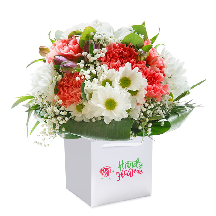 An expertly arranged bouquet rests gracefully in a squared white gift bag, its form and colour capturing the warm, welcoming character of Herne Hill. The bouquet centres on plush coral and peach carnations, their ruffled petals unfolding layer upon layer, and is enriched by vivid red carnations that anchor the design with vibrant energy. Interwoven among these, dainty white daisies with lively yellow-green eyes peek out, bringing a splash of brightness and optimism to the ensemble. Cascading through the blooms, sprigs of white baby's breath form airy bridges that infuse the bouquet with softness, while tightly held purple-tinged buds - perhaps alstroemeria not yet in full bloom - hint at beauty still to come. A halo of lush, shiny green foliage wraps the base, reminiscent of the tree-lined walkways along Herne Hill's Station Square, creating a strong yet elegant backdrop that frames the flowers above. The sturdy gift bag, adorned with twin ribbon handles, offers both functionality and refinement, ready to be carried away as a heartfelt present or delivered as a seamless surprise to a home off Norwood Road. The setting's pure white backdrop highlights every nuance, from the delicate textures of each bloom to the glossy detail of each leaf, and the precise rounding of the arrangement radiates a sense of careful preparation and abundance. Whether given for a joyous celebration or as a sincere gesture of affection, this bouquet captures a sense of freshness and elevated craftsmanship unique to Herne Hill's beloved floral scene.